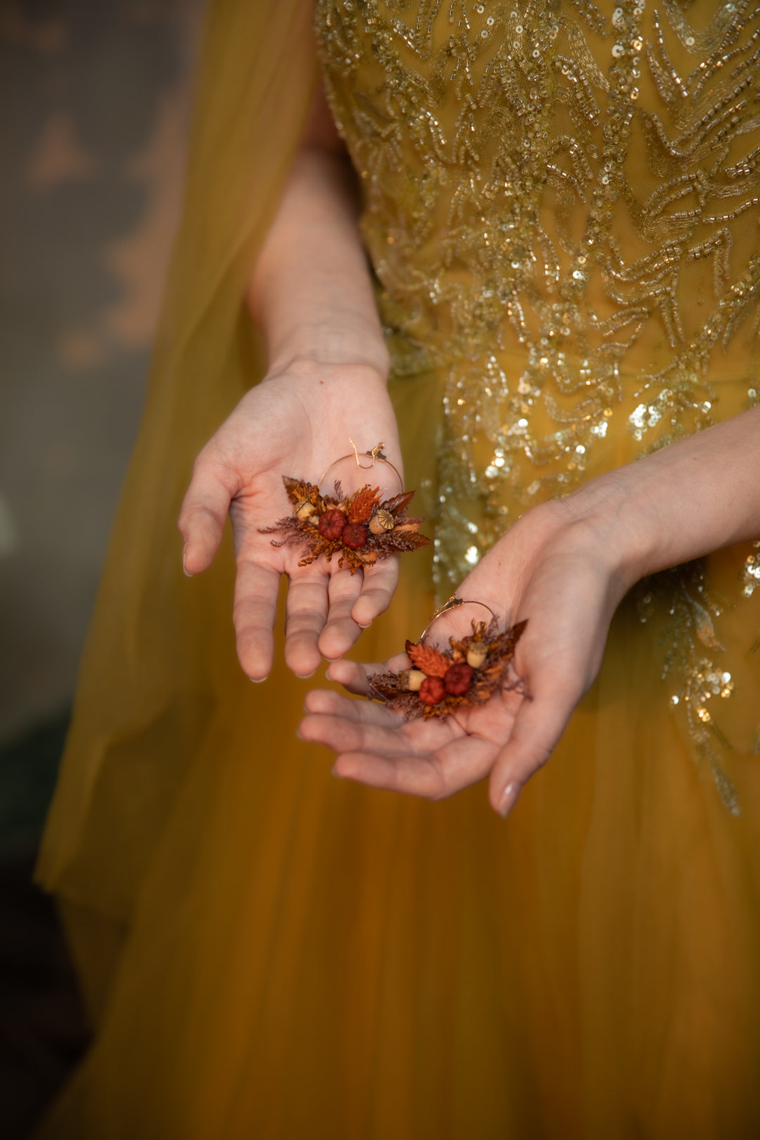 A close-up of a person wearing a shimmering gold sequined dress, holding two autumn earrings with pumpkins in their hands.