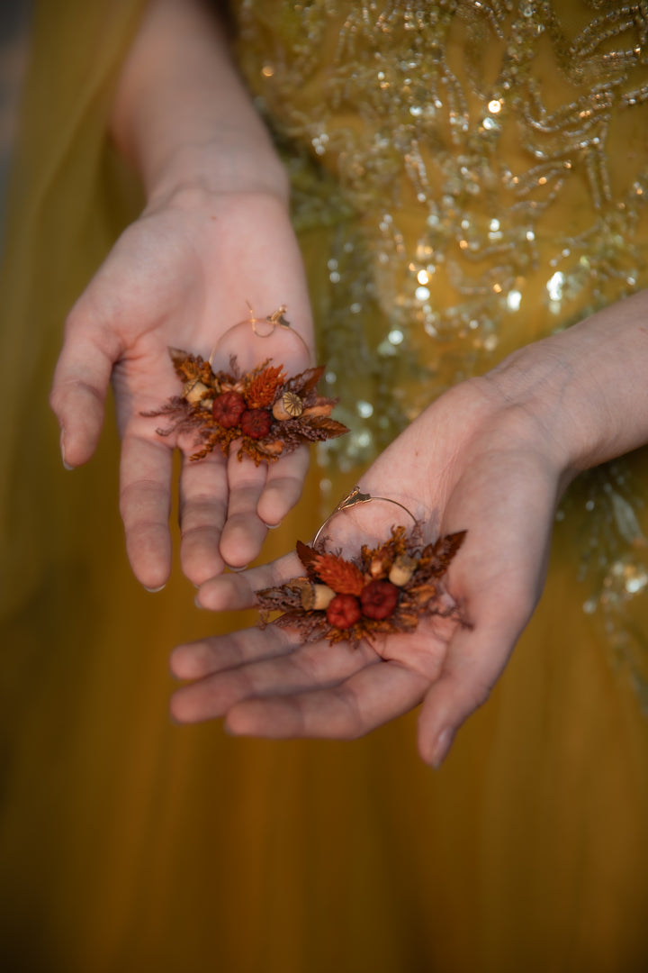 A close-up of a person's hands holding two small dried floral earrings  in the palm of their hands, wearing a shimmering gold dress against a warm golden background.