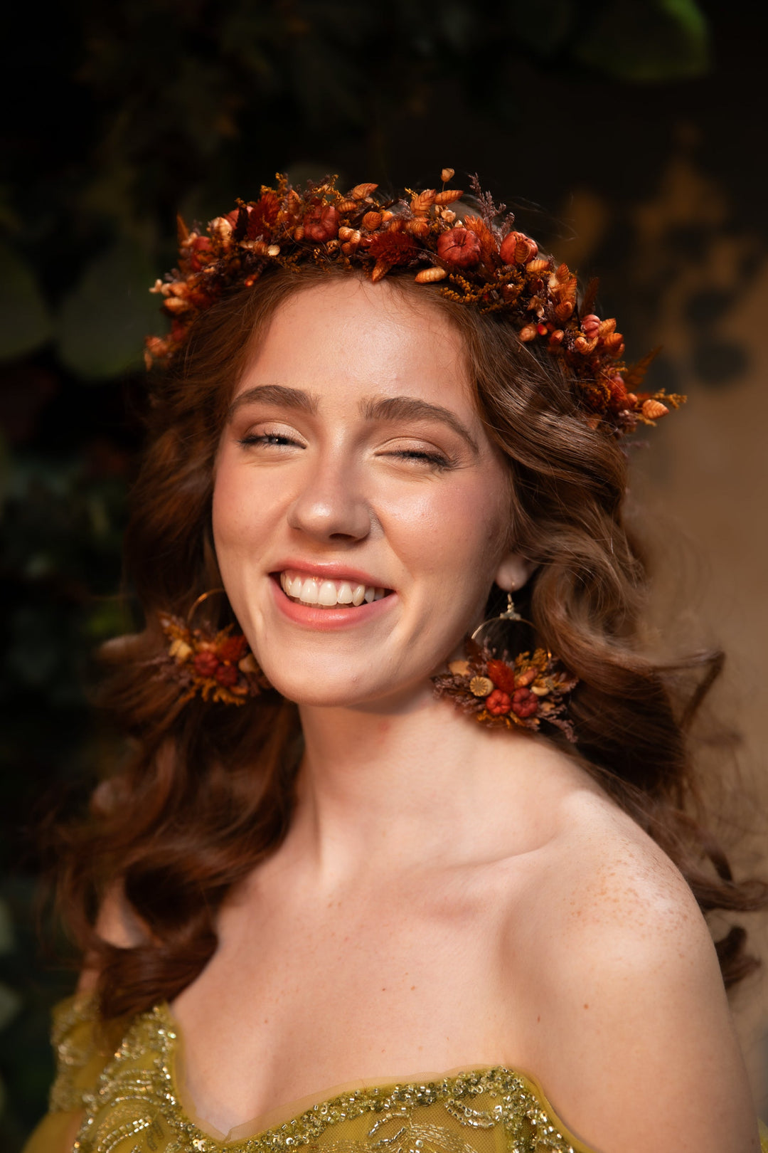 A smiling woman with wavy brown hair wearing an orange and brown floral crown and matching dangling earrings, with a gold embellished strapless top against a dark green foliage background.