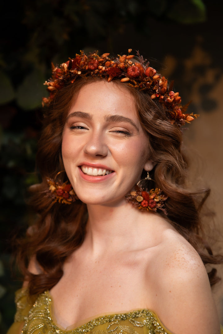 A smiling woman with wavy brown hair wearing an orange and brown floral crown and matching dangling earrings, with a gold embellished strapless top against a dark green foliage background.