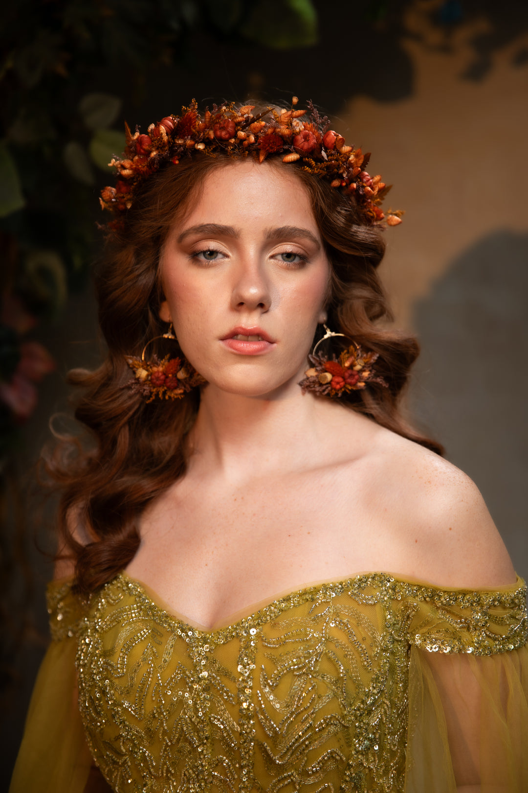 A woman wearing a warm olive-gold off-the-shoulder gown with intricate beadwork, a matching floral crown, and matching earrings, set against a dark, leafy background.