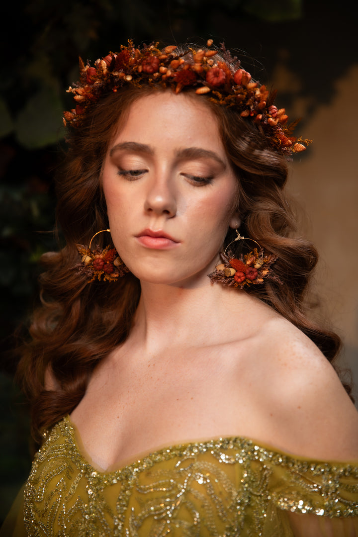 A woman with closed eyes wearing a floral crown in rust orange tones, large floral drop earrings, and an off-the-shoulder olive green sequined dress, set against a dark, soft-focus background.