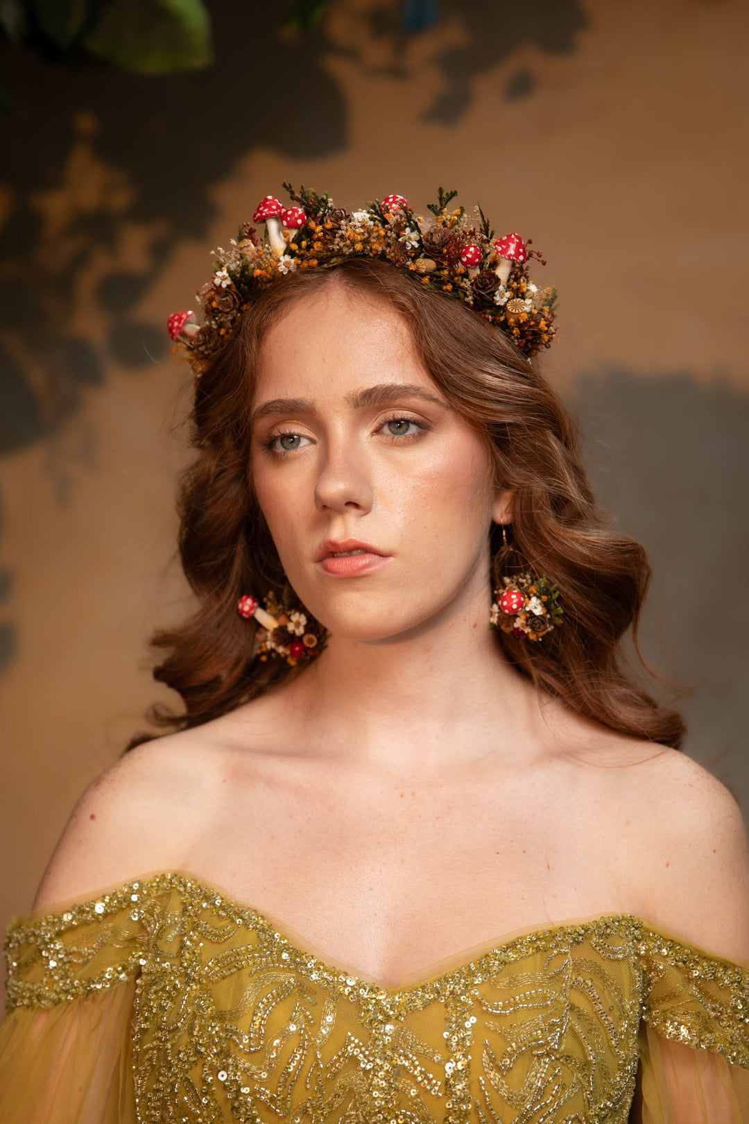 A woman with wavy brown hair wears a floral crown made of pink, red, and green flowers, paired with matching dangling earrings and an off-the-shoulder gold sequined gown against a warm brown background.
