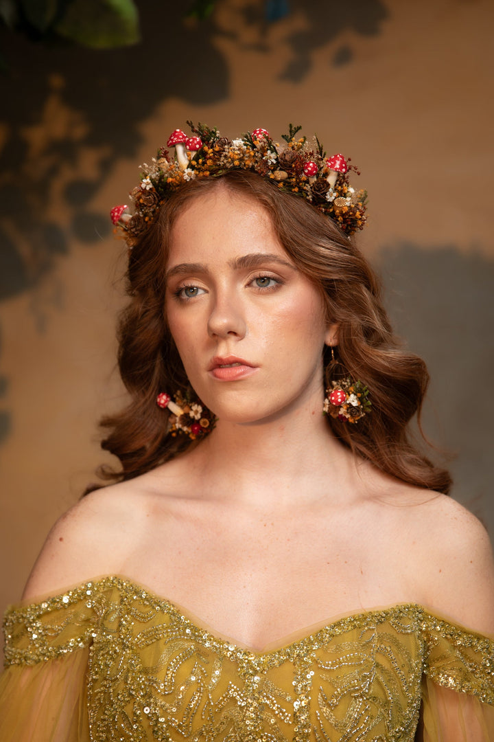 A woman with wavy brown hair wears a floral crown made of pink, red, and green flowers, paired with matching dangling earrings and an off-the-shoulder gold sequined gown against a warm brown background.
