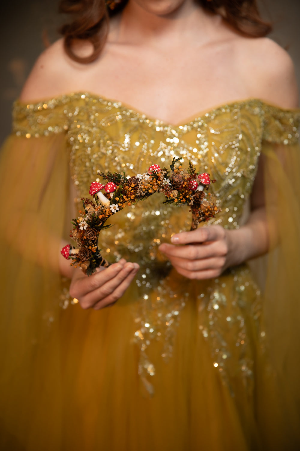 A woman wearing an off-the-shoulder gold sequined gown holds a decorative floral headband with mushrooms and small white flowers against a dark blurred background.