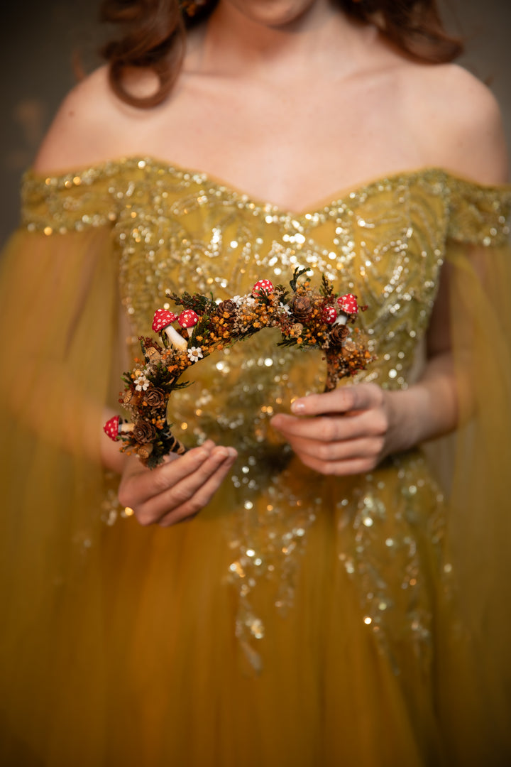 A woman wearing an off-the-shoulder gold sequined gown holds a decorative floral headband with mushrooms and small white flowers against a dark blurred background.