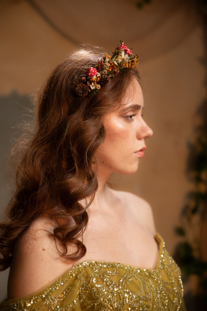 A woman in profile wearing a decorative floral crown with red mushrooms and a shimmering olive-gold off-the-shoulder dress, photographed in warm indoor lighting against a soft beige background.