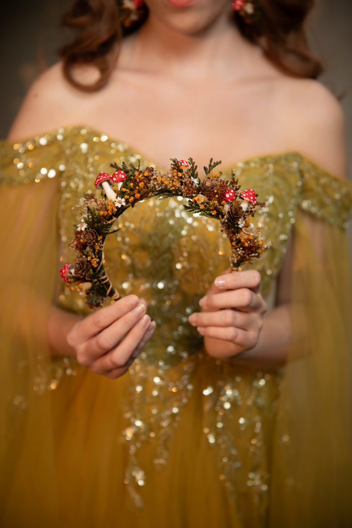 A woman in an off-the-shoulder gold sequined gown holds a floral crown wreath decorated with small red mushrooms , green leaves, and dried botanicals.