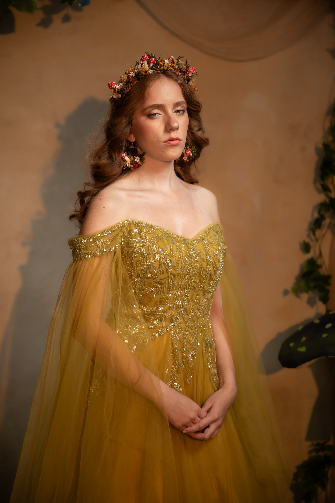 A woman wearing an off-the-shoulder golden yellow tulle gown with intricate gold sequin embroidery on the bodice, styled with a floral crown and matching earrings, set against a warm beige backdrop.