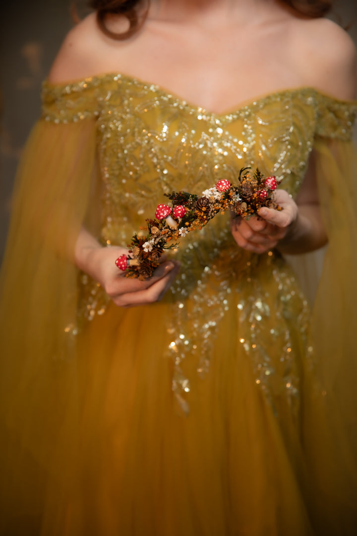 A woman in an off-the-shoulder gold tulle gown with sequined embellishments holds a dried floral crown featuring red mushrooms and brown leaves.