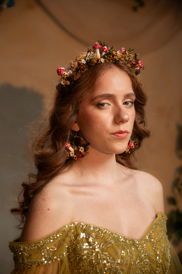 A woman with wavy brown hair wearing a floral crown and matching dangling earrings, dressed in an off-the-shoulder gold sequined gown against a warm beige background.