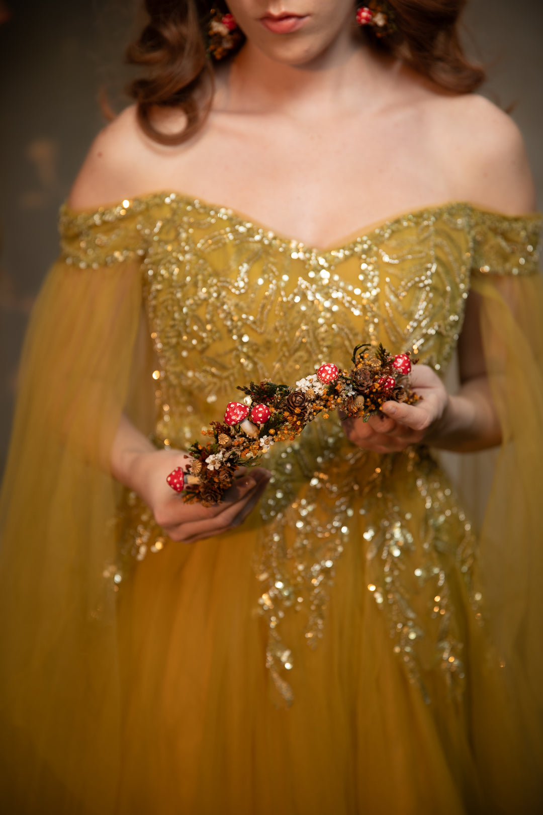 A woman wearing an off-the-shoulder gold sequined gown holds a floral headpiece with pink and gold flowers against a dark, softly lit background.