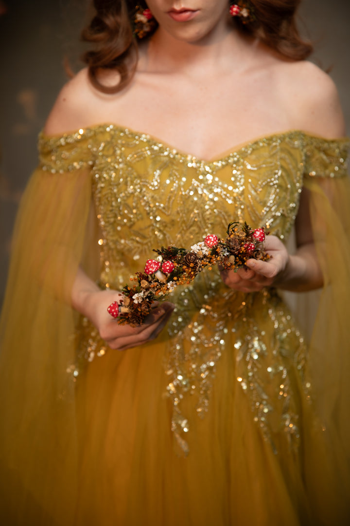 A woman wearing an off-the-shoulder gold sequined gown holds a floral headpiece with pink and gold flowers against a dark, softly lit background.
