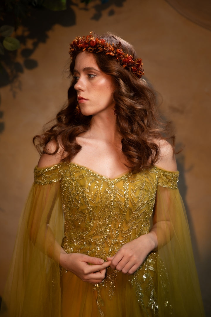 A woman wearing an ornate off-the-shoulder gown with intricate beadwork and sheer tulle sleeves, styled with a rust-orange floral crown against a warm brown studio backdrop.