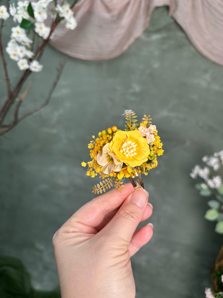 A hand holding a small flower hair clip with a bright yellow flower, smaller yellow buds, and beige leaves, set against a soft green textured background with blurred floral decorations.