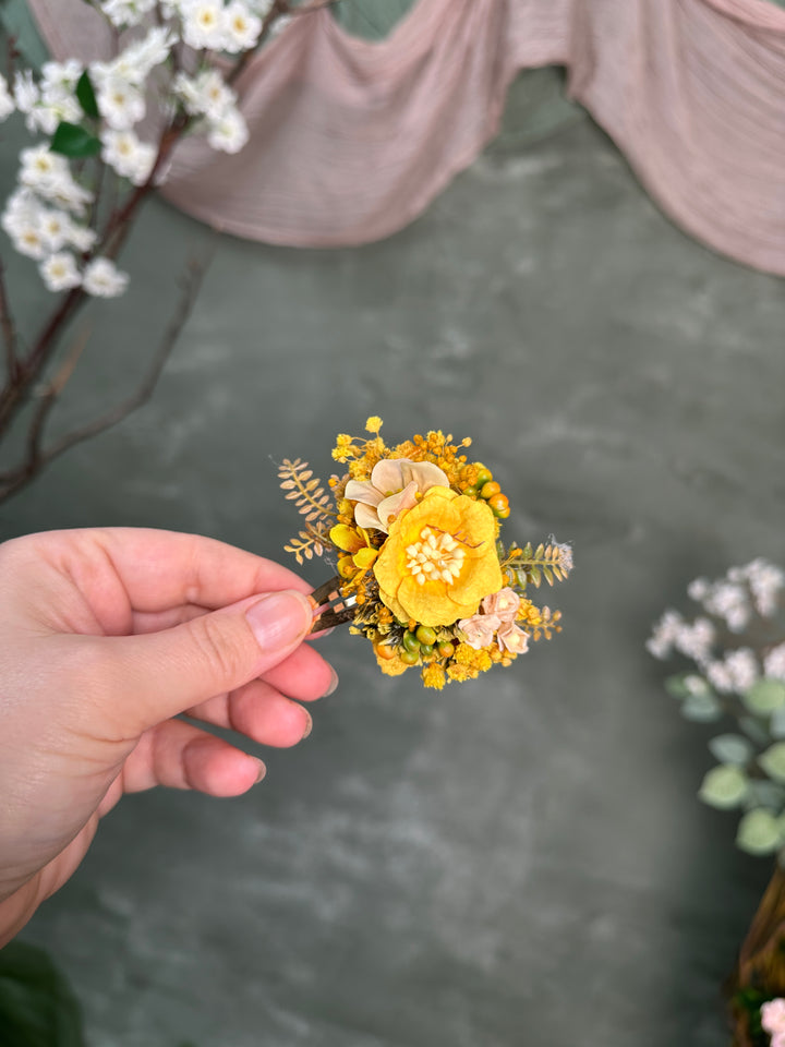 A hand holding a hair clip featuring a prominent yellow flower, smaller yellow blossoms, and pale peach and greenish foliage, set against a soft gray backdrop with blurred decorative floral elements.