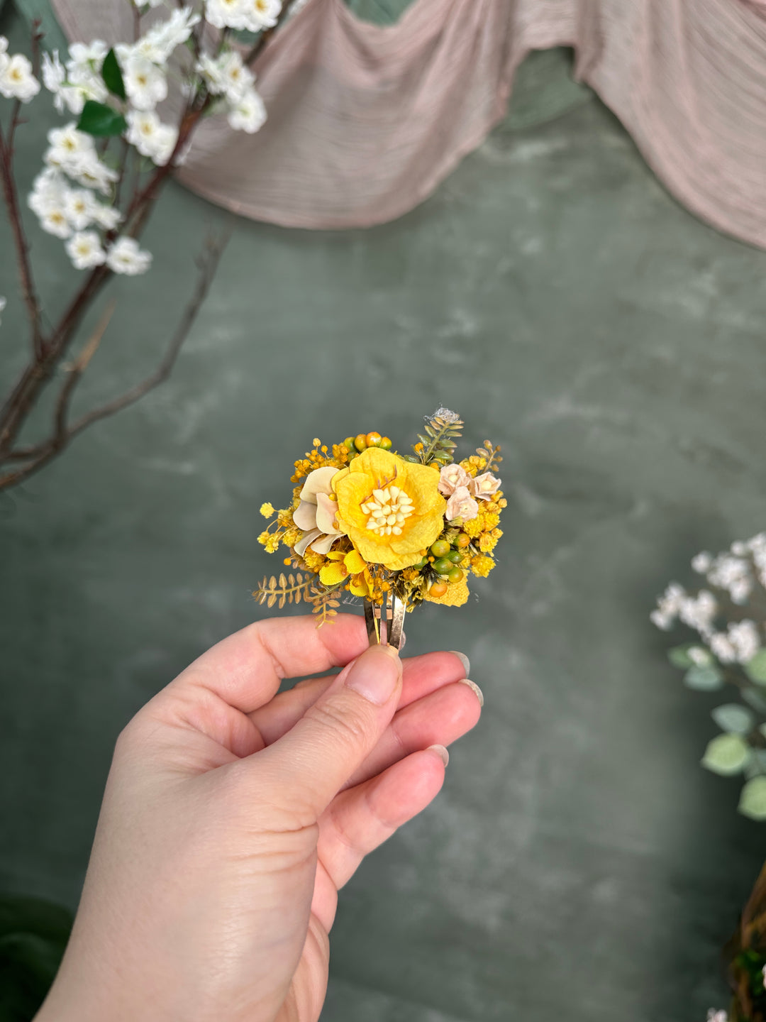 A hand holding a small flower hair clip with a bright yellow flower, smaller yellow blossoms, and ivory accents against a textured green backdrop.