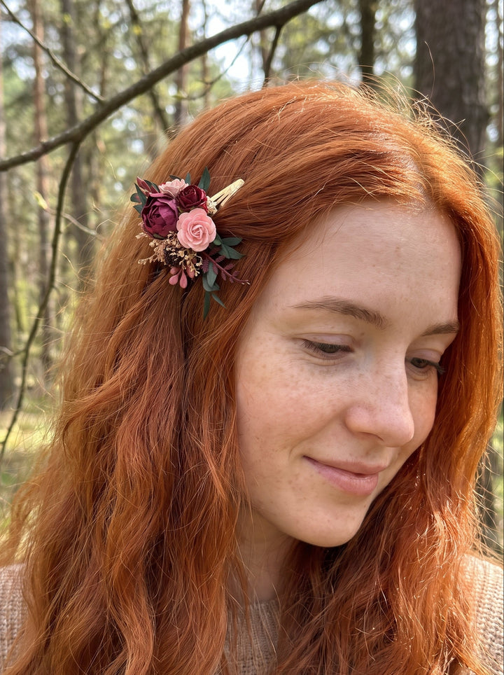 A woman with long, wavy red hair wearing a small floral hair clip with pink, burgundy, and green flowers, photographed outdoors in a forest setting.