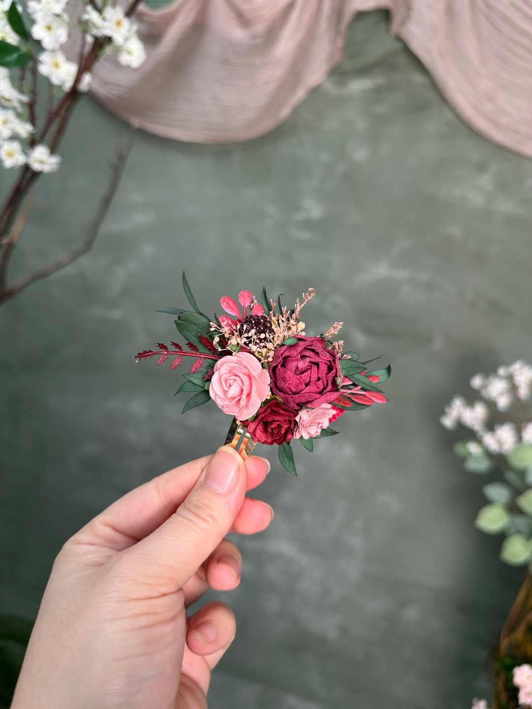 A hand holding a small artificial floral snap clip with pink and burgundy roses, green leaves, and dried botanicals against a soft gray-green backdrop.