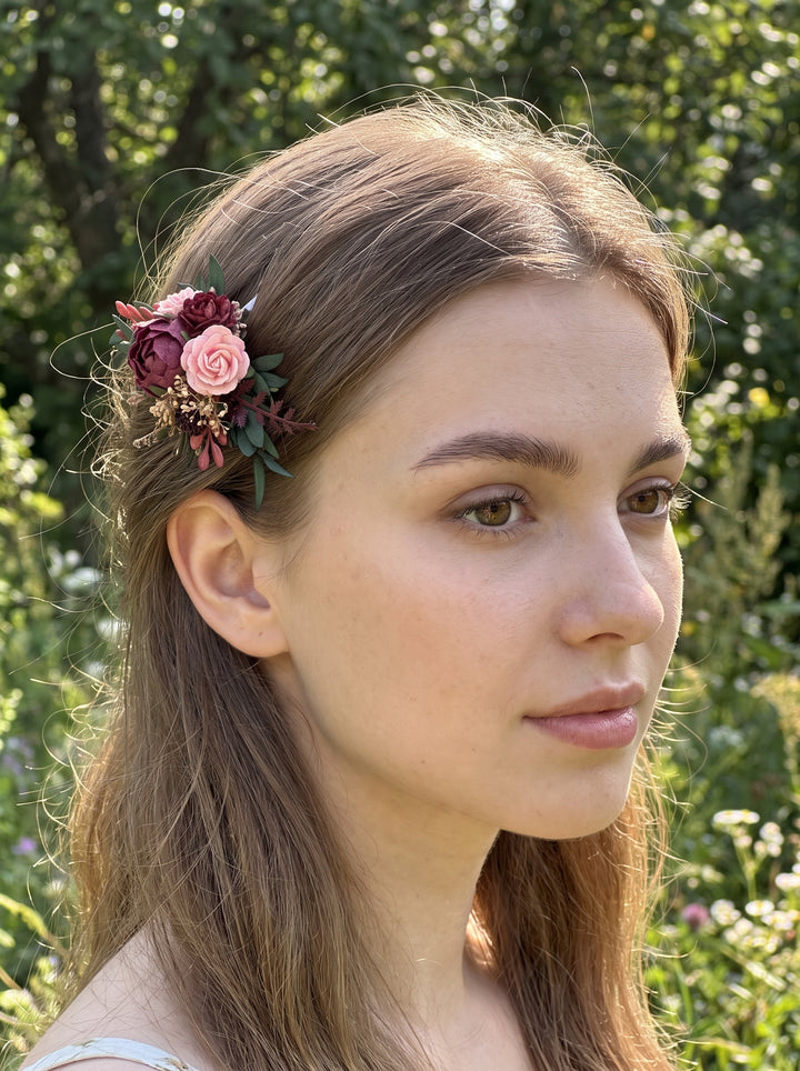 A woman outdoors wearing a floral hair accessory with pink and burgundy flowers and green leaves tucked into her brown hair, with a blurred green garden background.