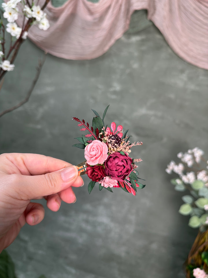 A hand holding a small artificial floral hair accessory with pink and burgundy roses, green leaves, and dried filler flowers against a soft gray backdrop with draped fabric and blurred flowers.