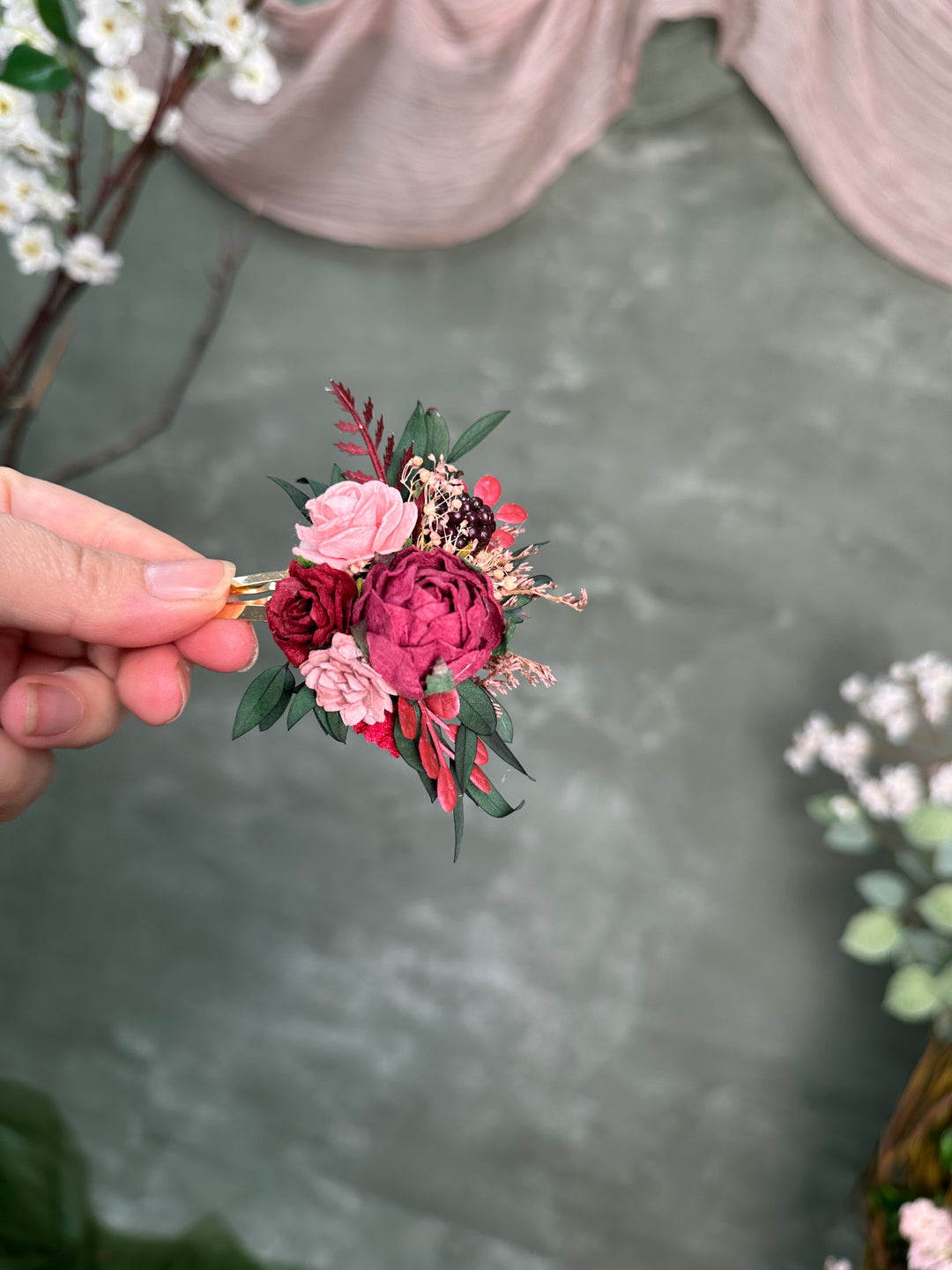 A hand holding a small artificial floral snap clip with pink and burgundy flowers, green leaves, and a gold-tone clip against a soft gray-green backdrop with blurred decorative flowers.