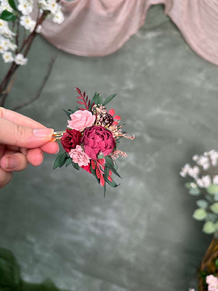 A hand holding a small artificial floral snap clip with pink and burgundy flowers, green leaves, and a gold-tone clip against a soft gray-green backdrop with blurred decorative flowers.
