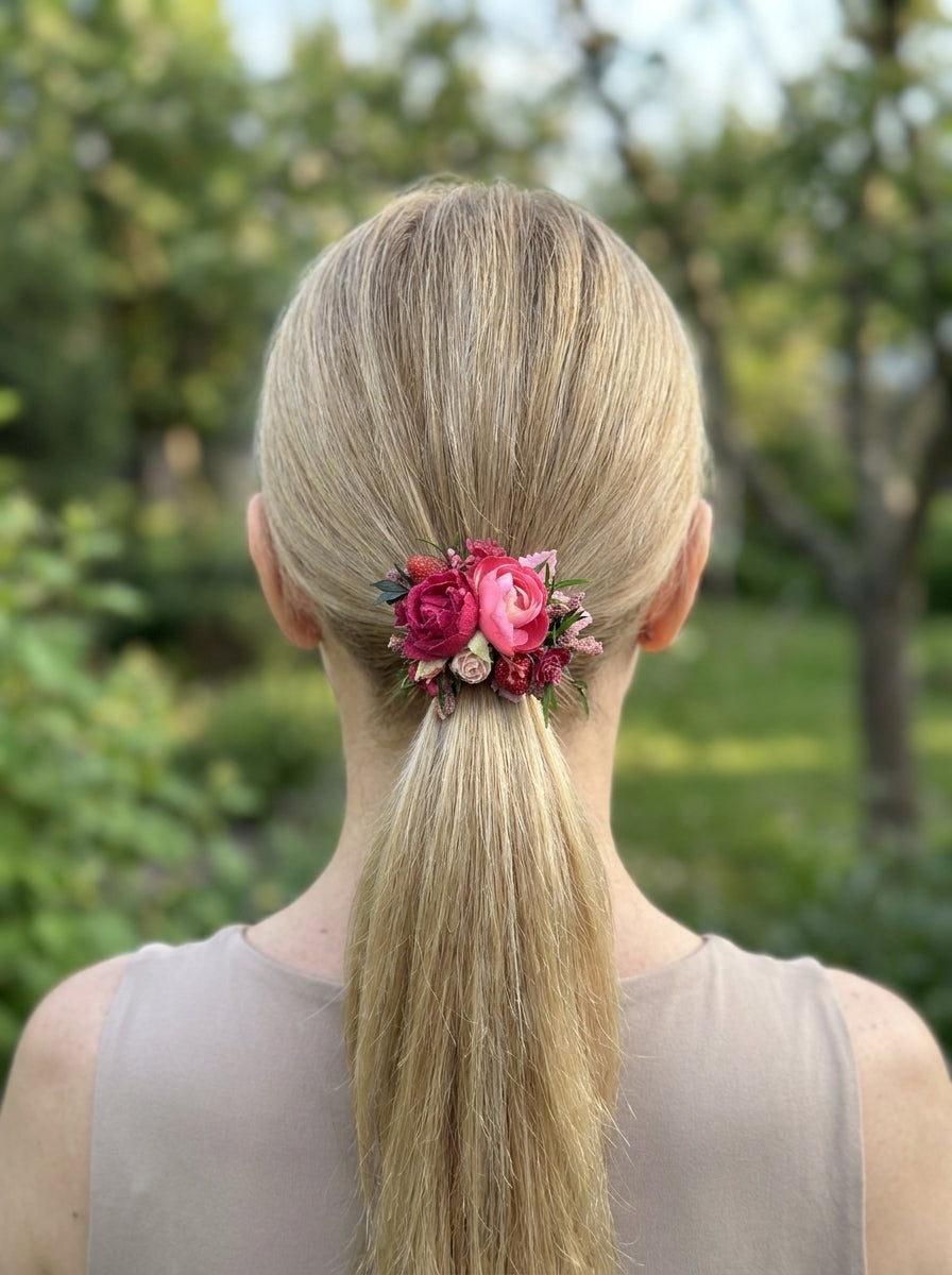 A blonde woman viewed from behind, wearing a pink and red floral hair accessory in a ponytail, with a blurred green outdoor background.