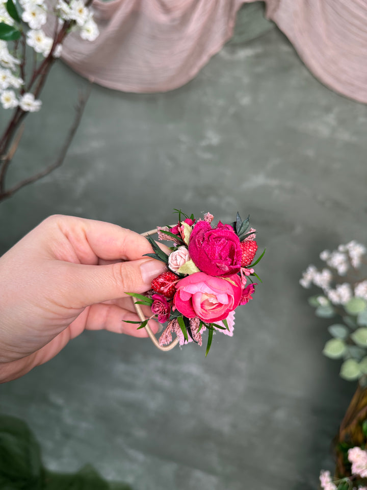 A hand holding a small floral hair accessory with bright pink and red flowers, green leaves, and a pink ribbon, set against a soft gray background with blurred decorative flowers.