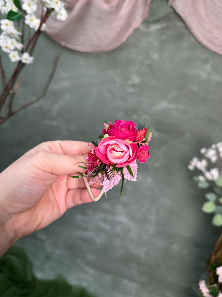 A hand holding a small pink floral hair tie with a pink rose, smaller pink blossoms, and green leaves against a soft gray-green backdrop with blurred decorative flowers.