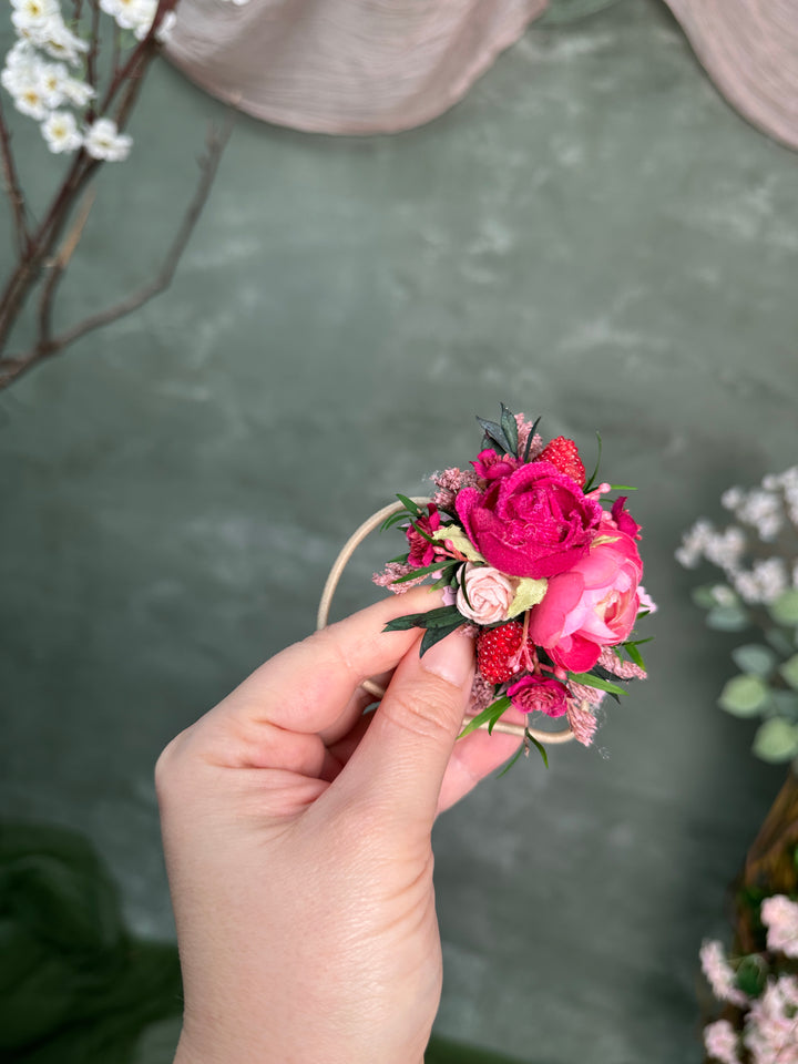 A hand holding a small floral wrist corsage featuring vibrant pink and red roses, berries, and green leaves, set against a textured green backdrop with blurred decorative elements.