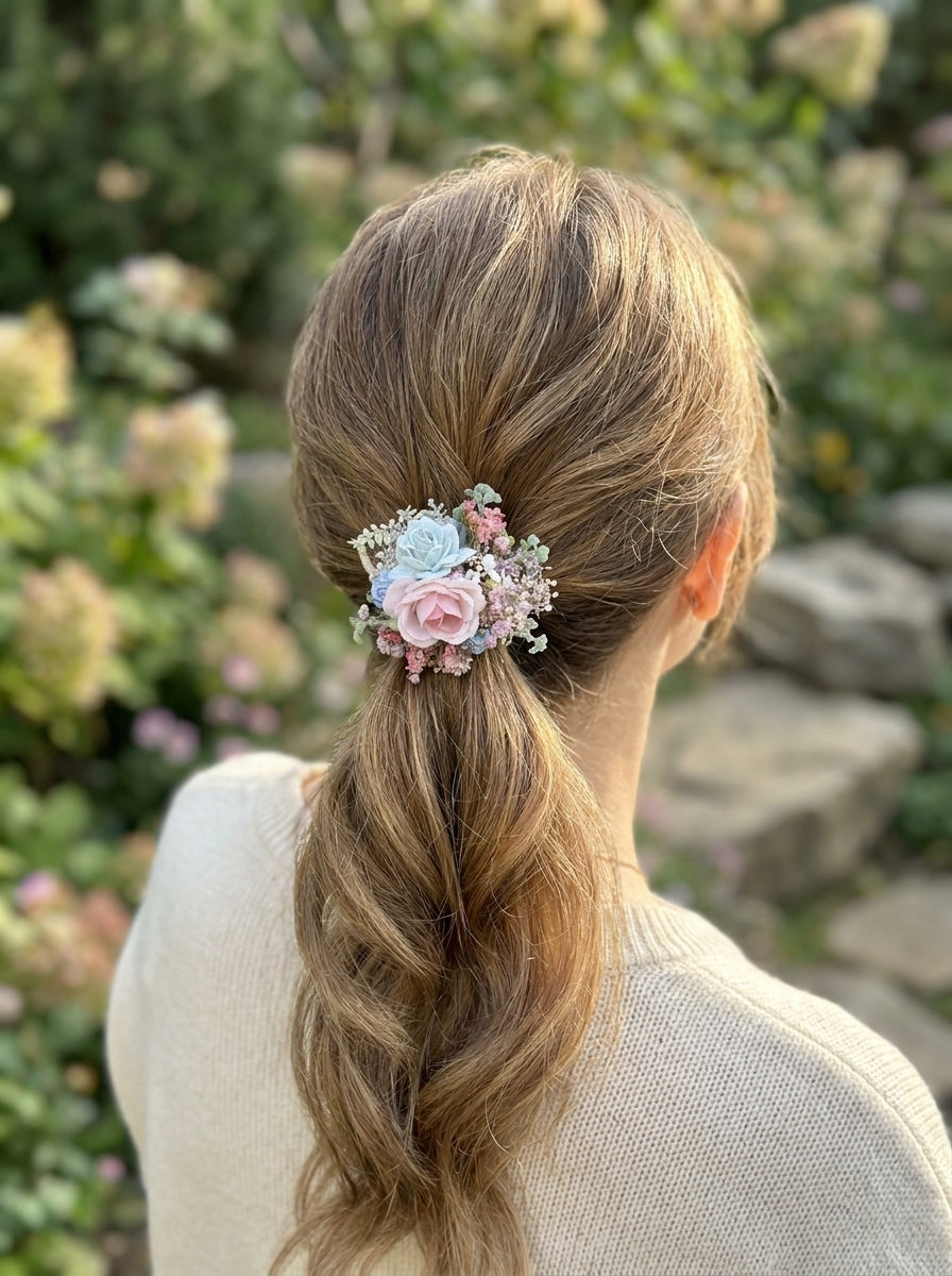 A woman viewed from behind with her hair in a low ponytail, wearing a small floral hair accessory with pink and light blue flowers in a garden setting.