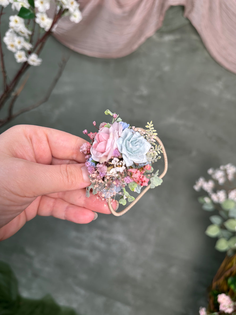 A hand holding a small floral hair accessory with pink and light blue faux roses, delicate green leaves, and tiny purple flowers attached to a thin beige band.