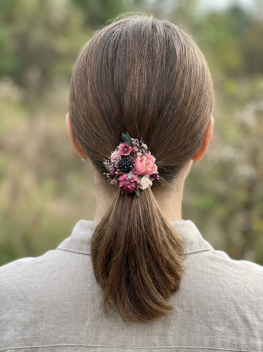 A woman viewed from behind wearing a hair accessory with pink and burgundy flowers and green leaves, holding her hair in a low ponytail. She is wearing a light beige linen shirt against a blurred outdoor background.
