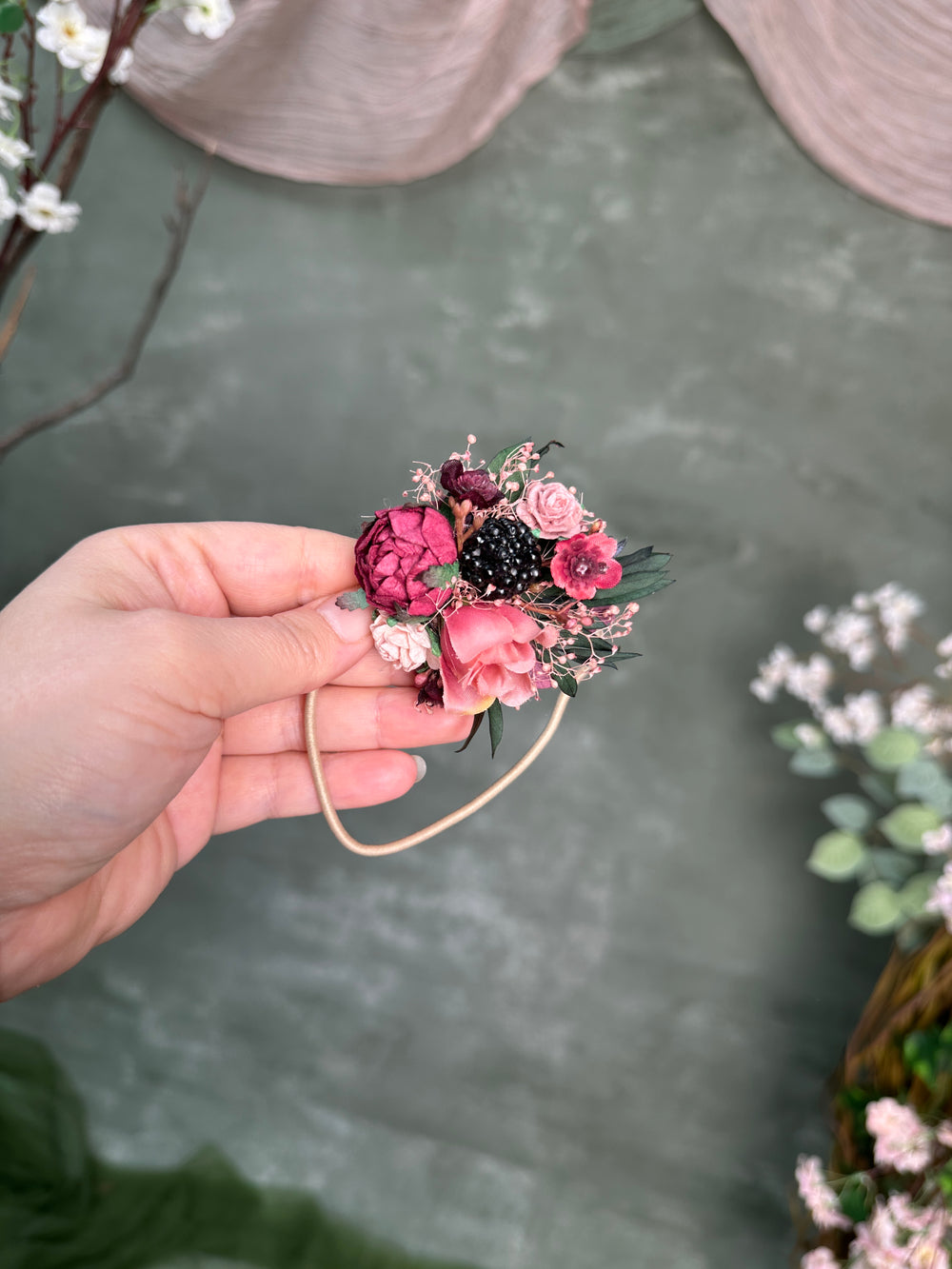 A hand holding a small floral hair accessory with pink and burgundy flowers, green leaves, and a beige elastic band, set against a textured gray-green background with blurred decorative flowers.