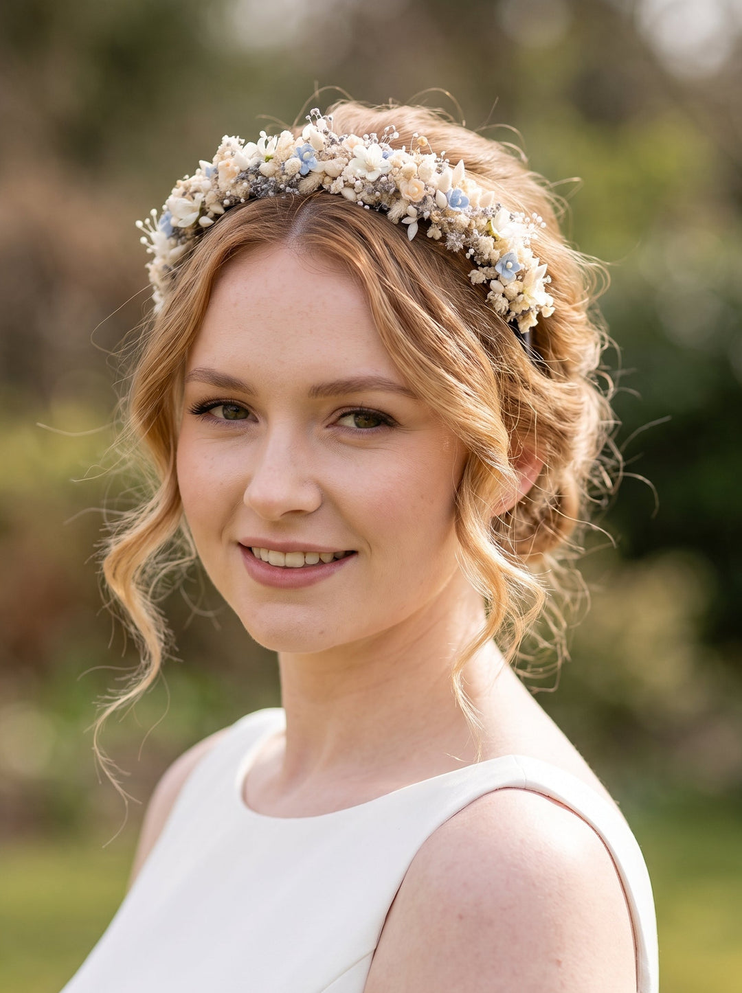 A woman outdoors wearing a delicate floral headpiece crown with small cream, white, and pale blue flowers, paired with a white sleeveless dress.