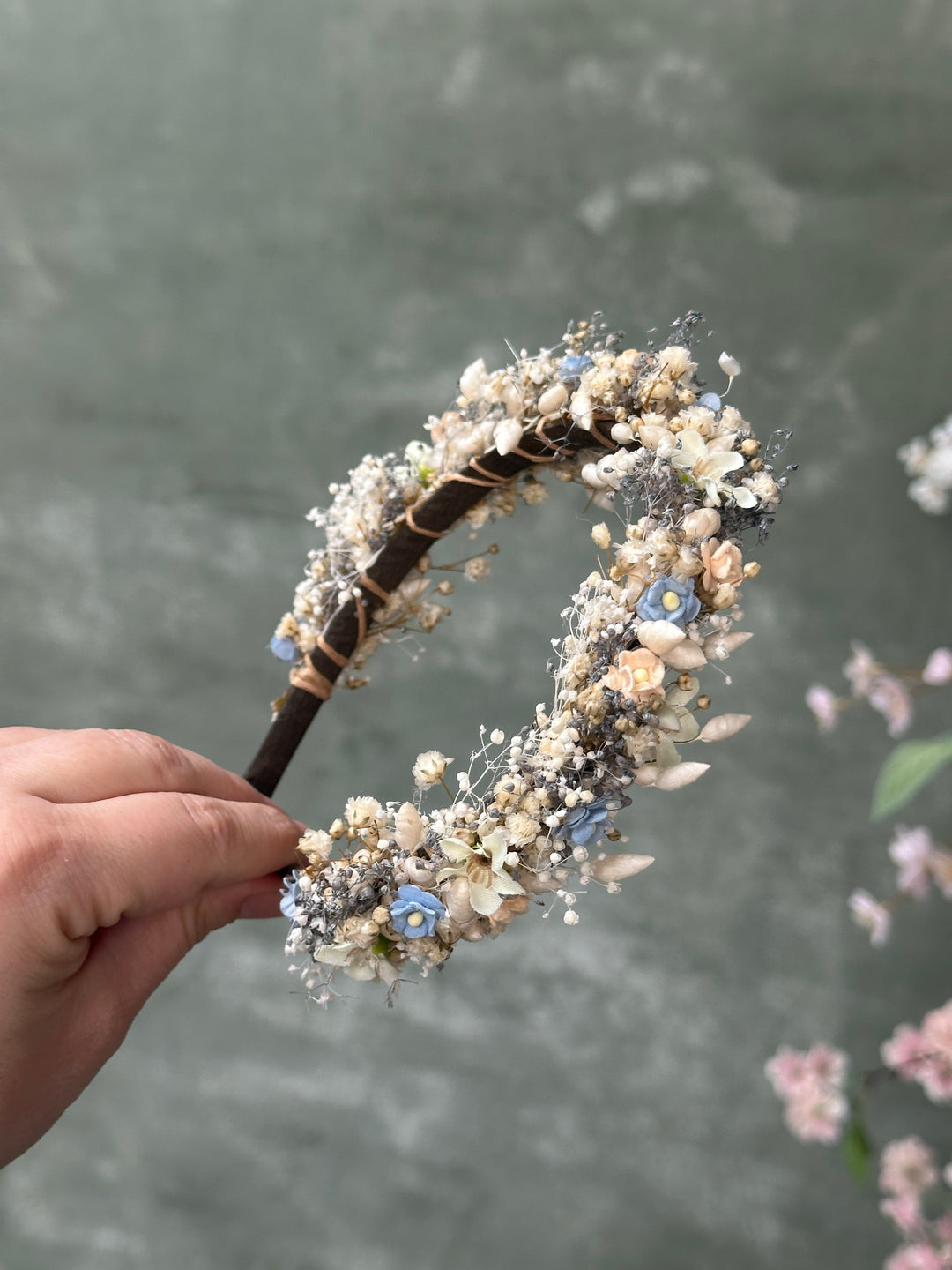 A hand holding a decorative headband adorned with white and beige flowers, small blue blossoms, and brown twine wrapping, set against a soft gray textured background.
