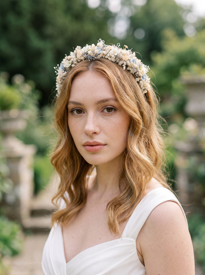 A woman with wavy light brown hair wearing a delicate floral crown headband and a white dress, photographed outdoors in a softly blurred garden setting.