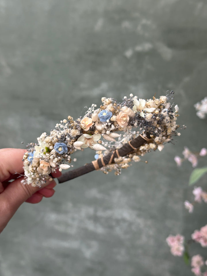 A hand holding a decorative headband adorned with flowers, small shells, and blue beads against a soft gray textured background.