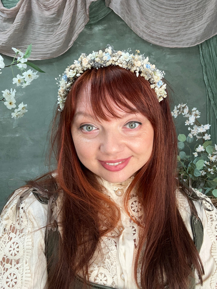 A woman with long auburn hair and a white floral crown lying on a dark green textured blanket, wearing a cream lace top.
