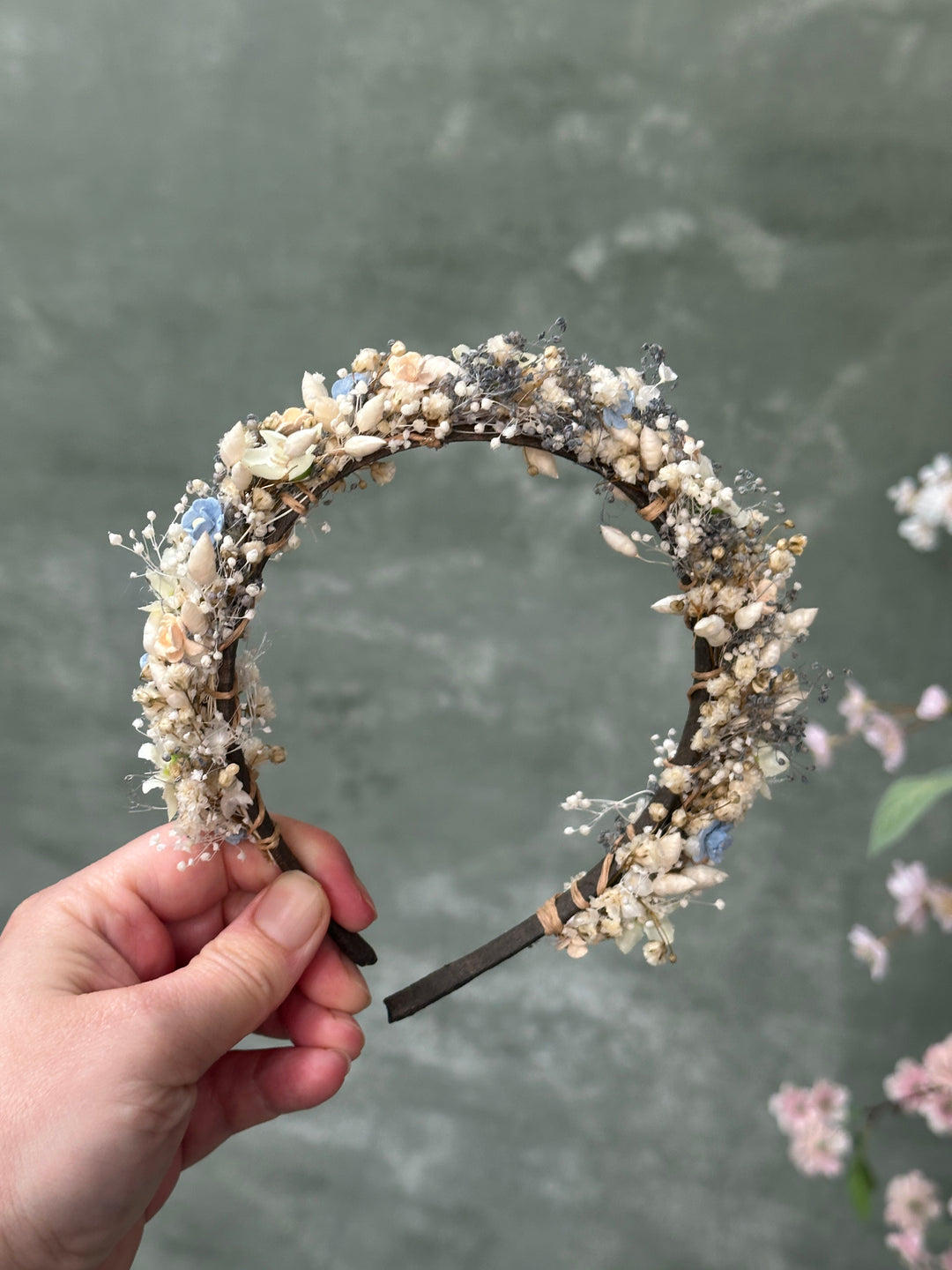 A handmade circular headband decorated with flowers held by a hand against a textured gray background.