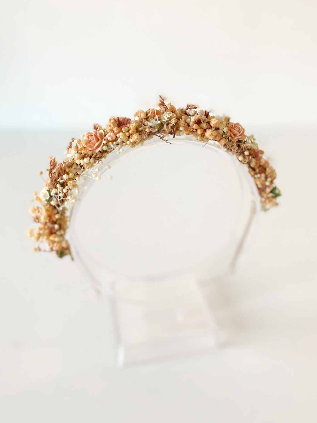 A floral crown with muted peach, beige, and brown botanicals displayed on a clear acrylic stand against a soft white background.