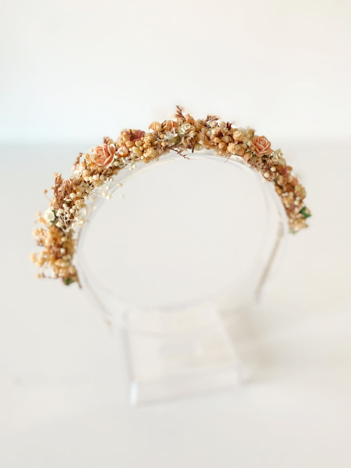A floral crown with muted peach, beige, and brown botanicals displayed on a clear acrylic stand against a soft white background.