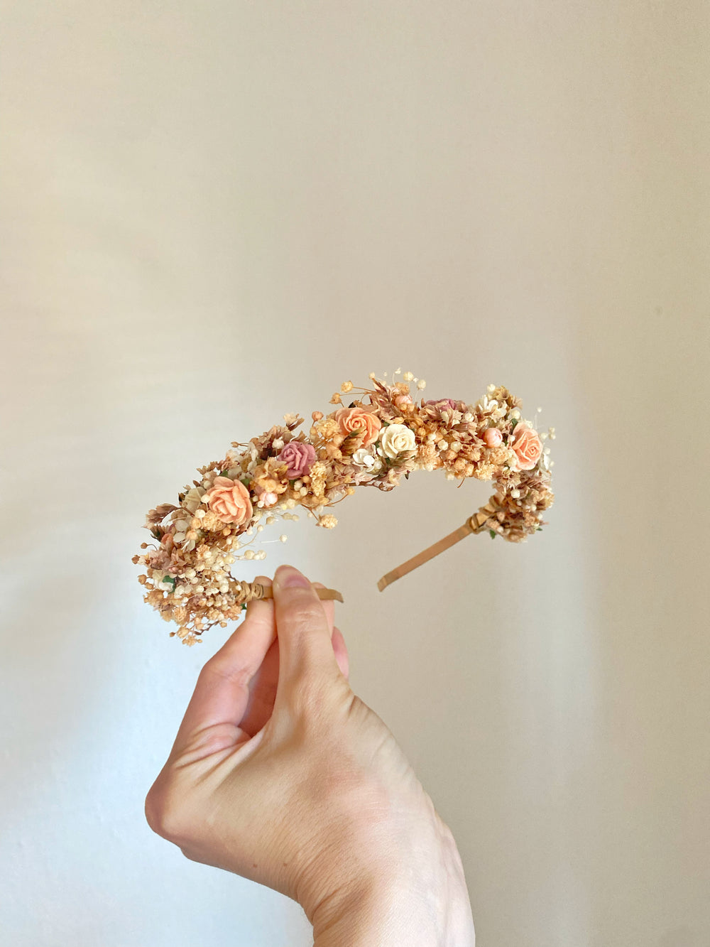 A hand holding a delicate floral headband with small peach, pink, and cream flowers on a light beige background.