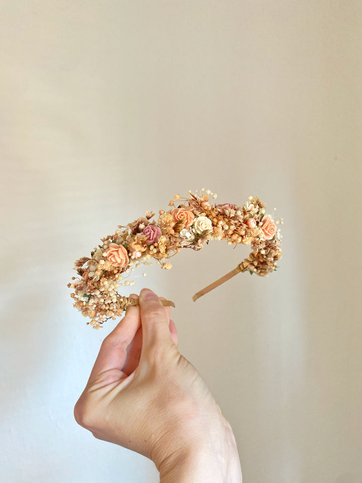 A hand holding a delicate floral headband with small peach, pink, and cream flowers on a light beige background.
