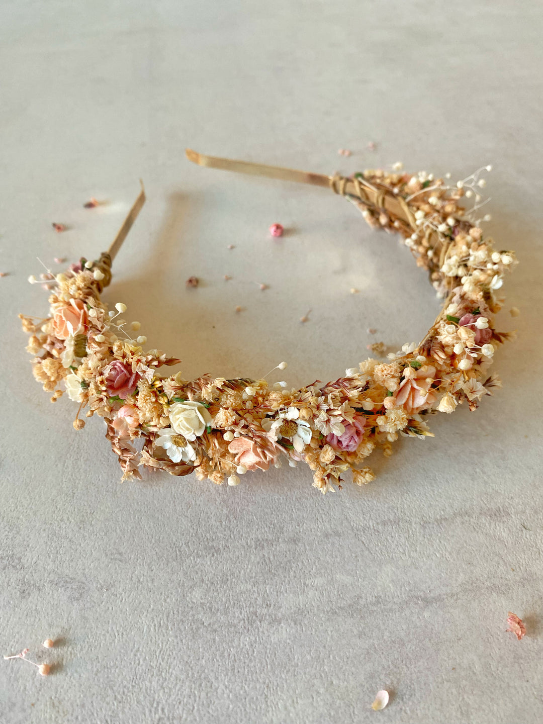 A delicate floral headband featuring flowers in soft peach, pink, cream, and muted green tones, arranged on a light gray textured surface.