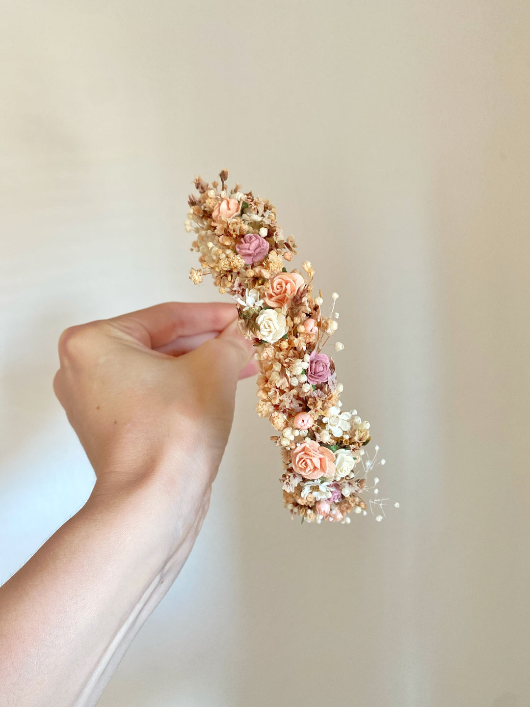 A hand holding a delicate flower hair accessory featuring small peach, pink, and cream roses with gold-toned glitter and tiny white filler flowers, set against a soft beige background.