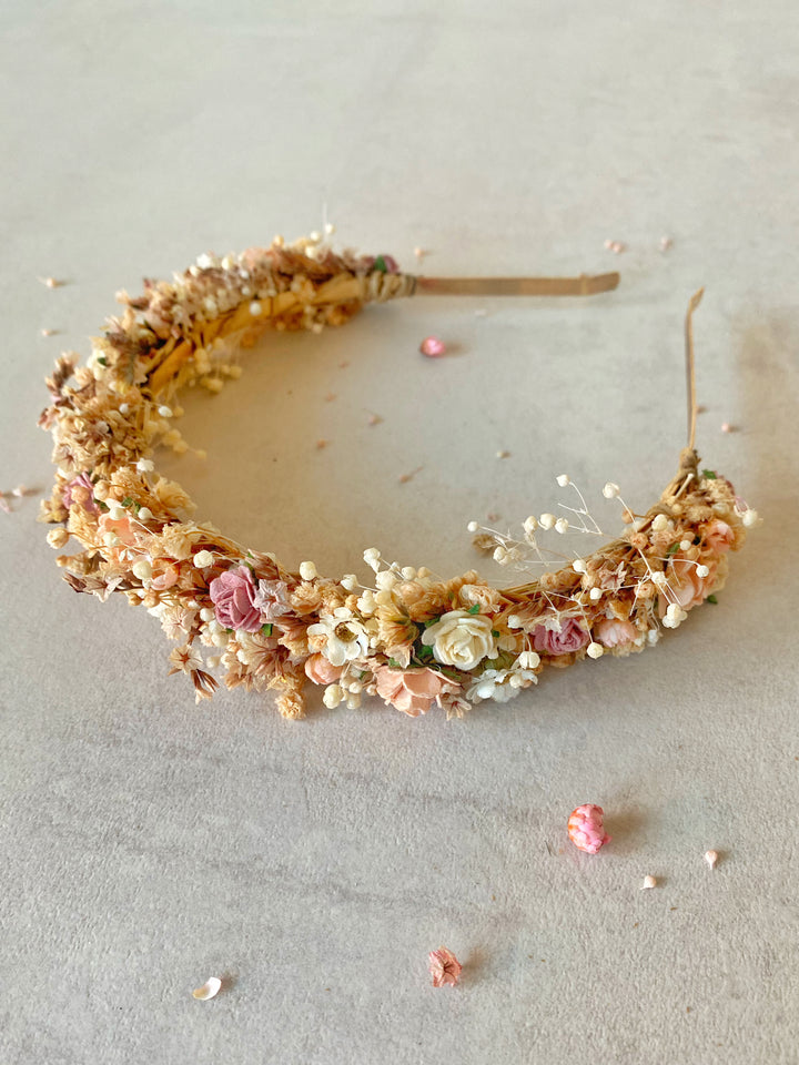 A delicate floral headband decorated with flowers, baby's breath, and small faux blossoms in soft pink, cream, and tan tones, displayed on a neutral light gray surface.