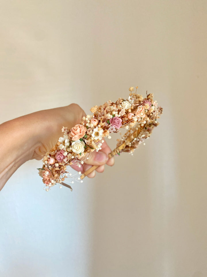 A hand holding a delicate flower headband with small faux roses, tiny white flowers, and pearl-like beads on a neutral beige background.