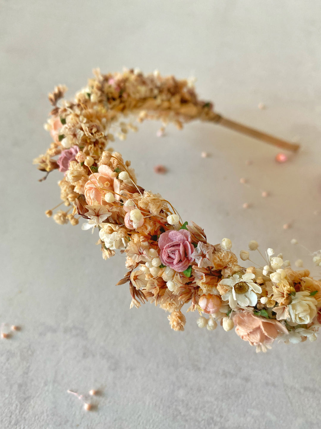 A floral headband featuring small pink and peach roses, cream blossoms, and delicate sprigs on a light beige background.